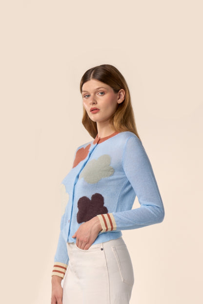 Cardigan bleu ciel en maille, manches longues, motifs nuages multicolores et poignets rayés, porté avec un pantalon écru — Pote aux Roses Paris